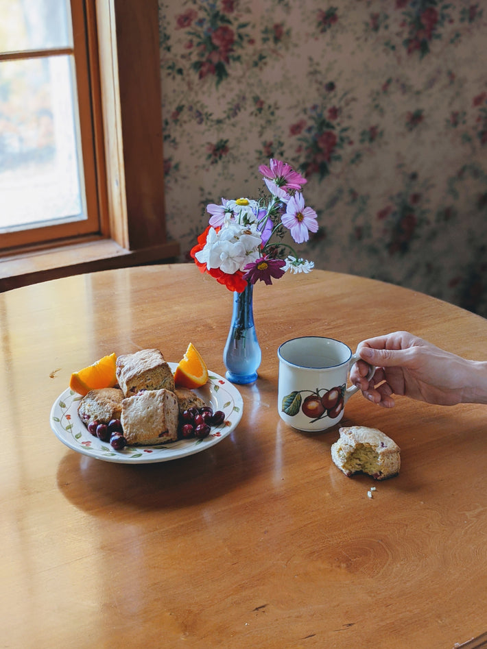 Orange-cranberry scones and grapes on a plate with orange wedges on a wooden table; mug beside a bitten scone and a blue vase of flowers, hand holding mug.