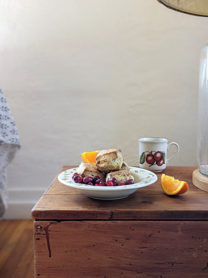 A plate of orange cranberry biscuits with grapes and an orange slice on a wooden table, next to a cup.