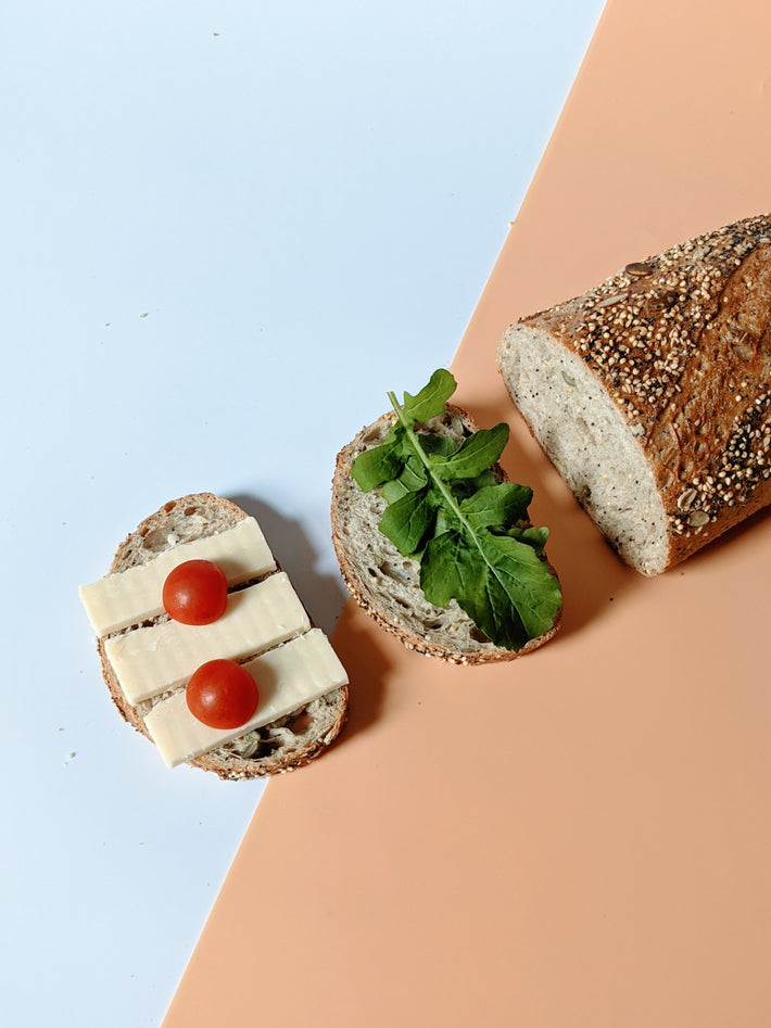 Slice of bread topped with cheese, cherry tomatoes, and arugula, with a loaf of bread beside it.