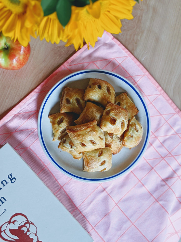 A plate of apple pie bites on a pink tablecloth with flowers and an open book nearby.