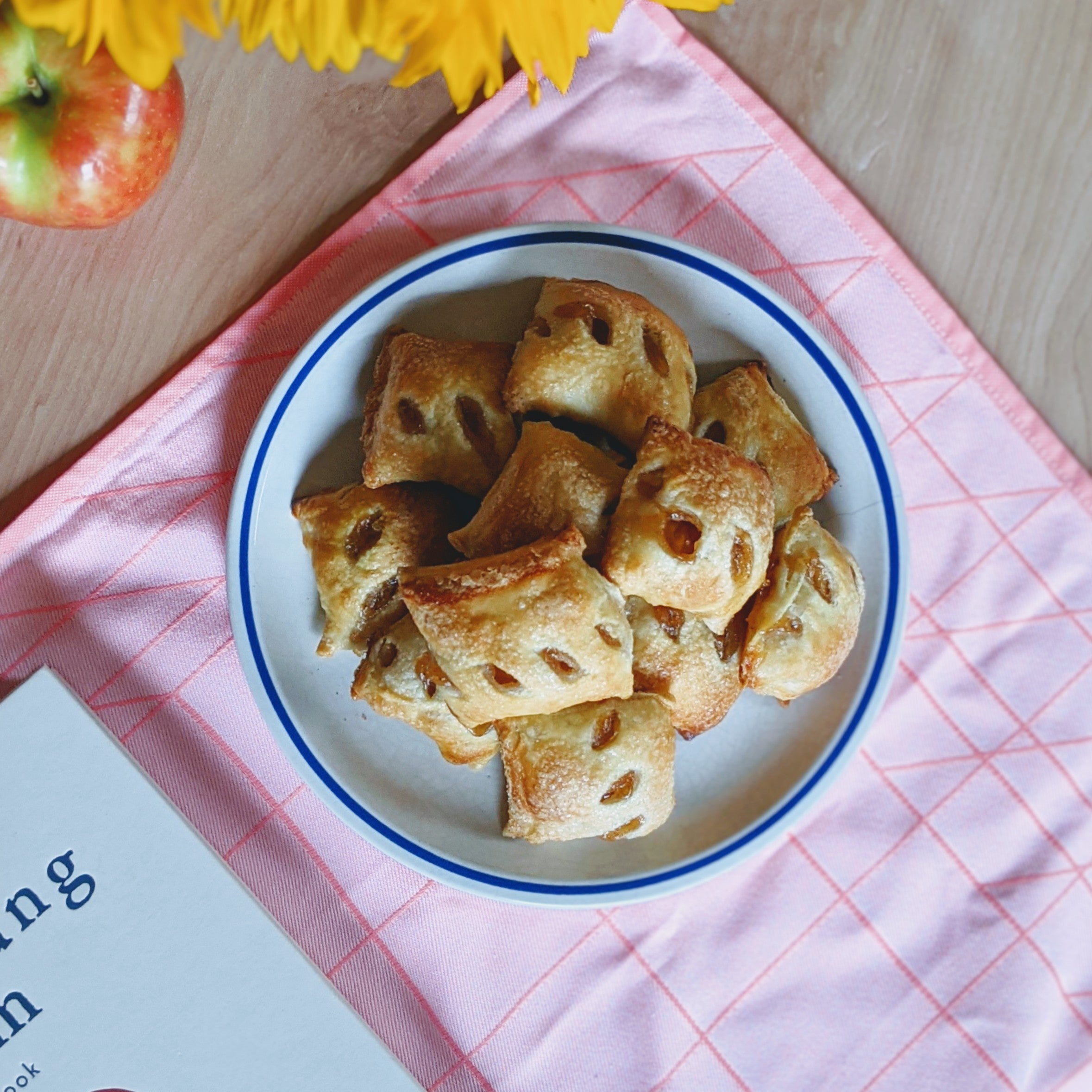 Puff pastries with raisin slits on a plate over a pink checked napkin; apple and sunflowers nearby, visible text 'ing'.