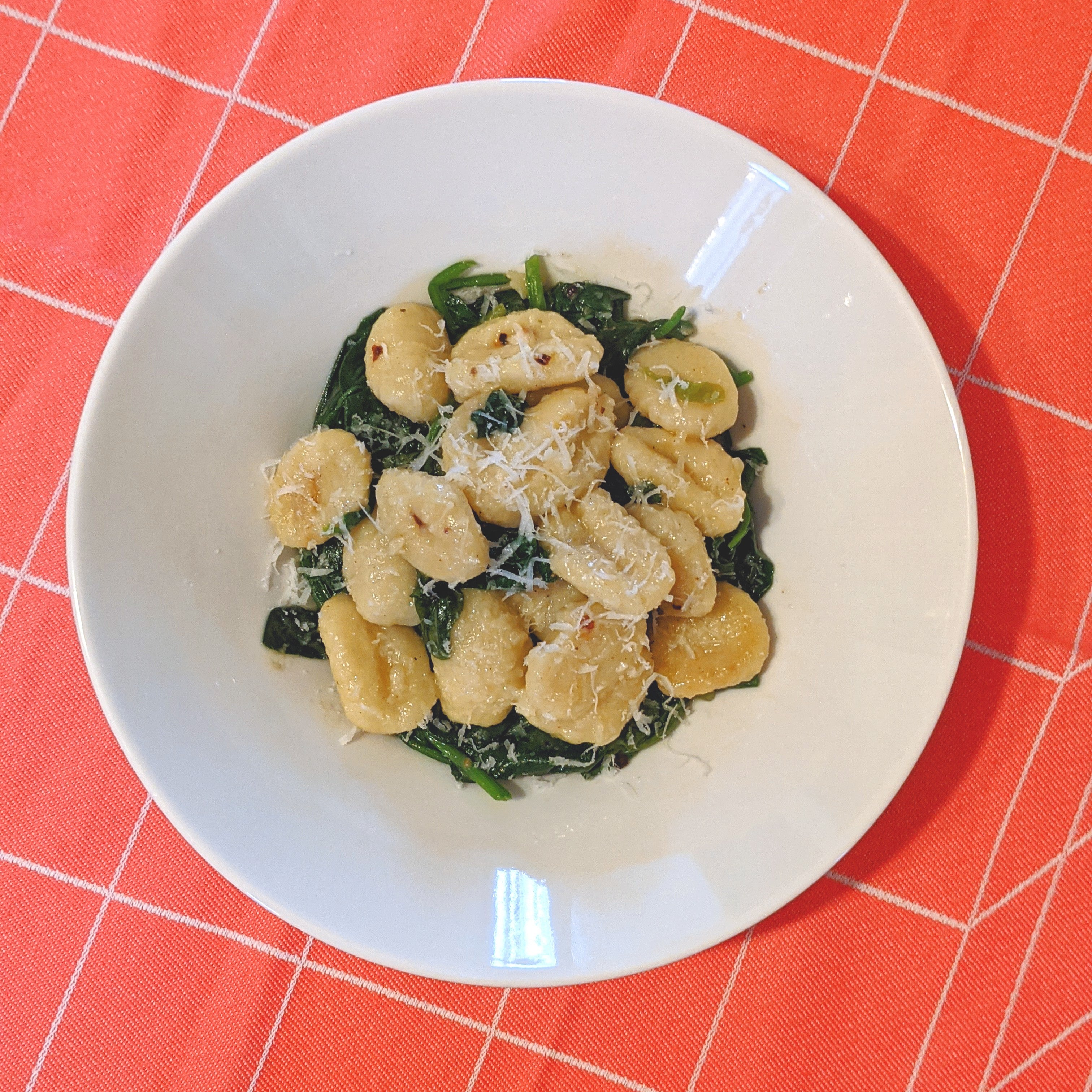 Bowl of gnocchi with sauteed spinach and grated Parmesan on a white plate over an orange checkered tablecloth.