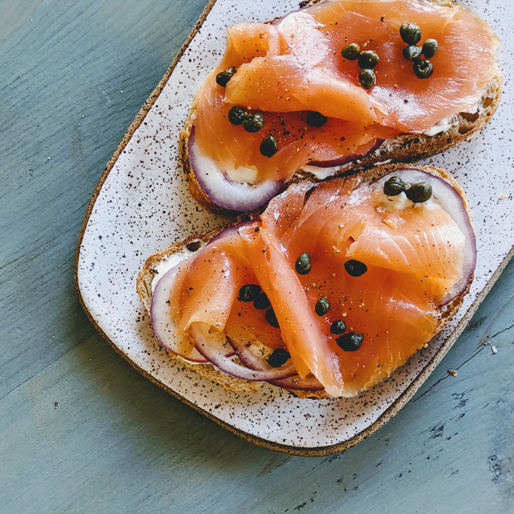 Two open-faced toasts with smoked salmon, red onion, cream cheese and capers on a speckled oval plate.