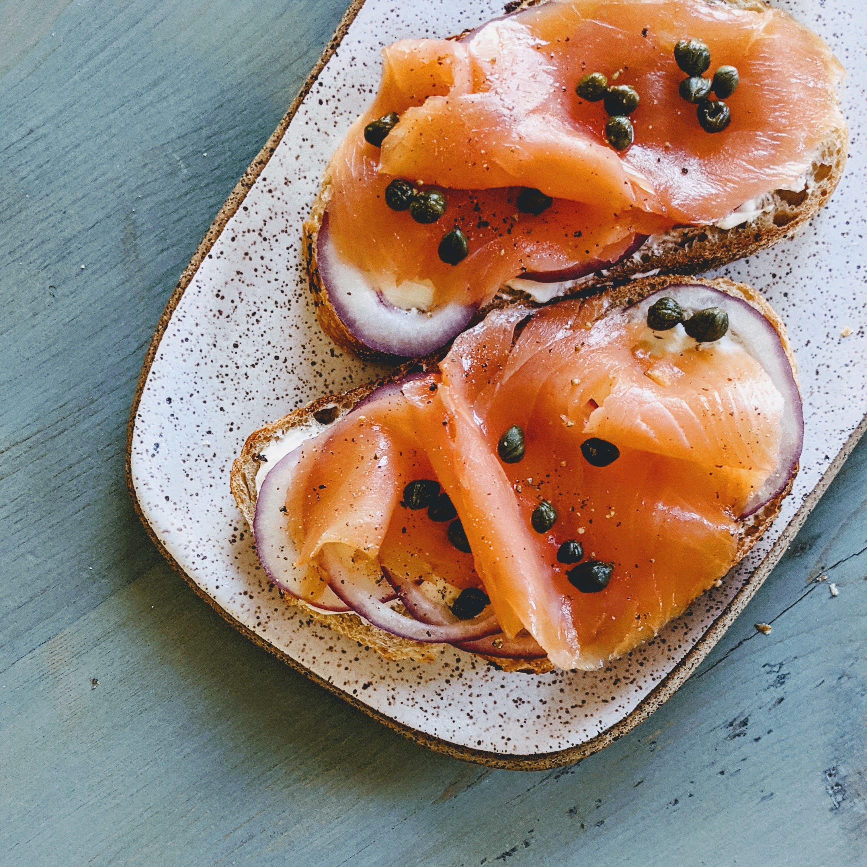 Two open-faced toasts with smoked salmon, red onion, cream cheese and capers on a speckled oval plate.