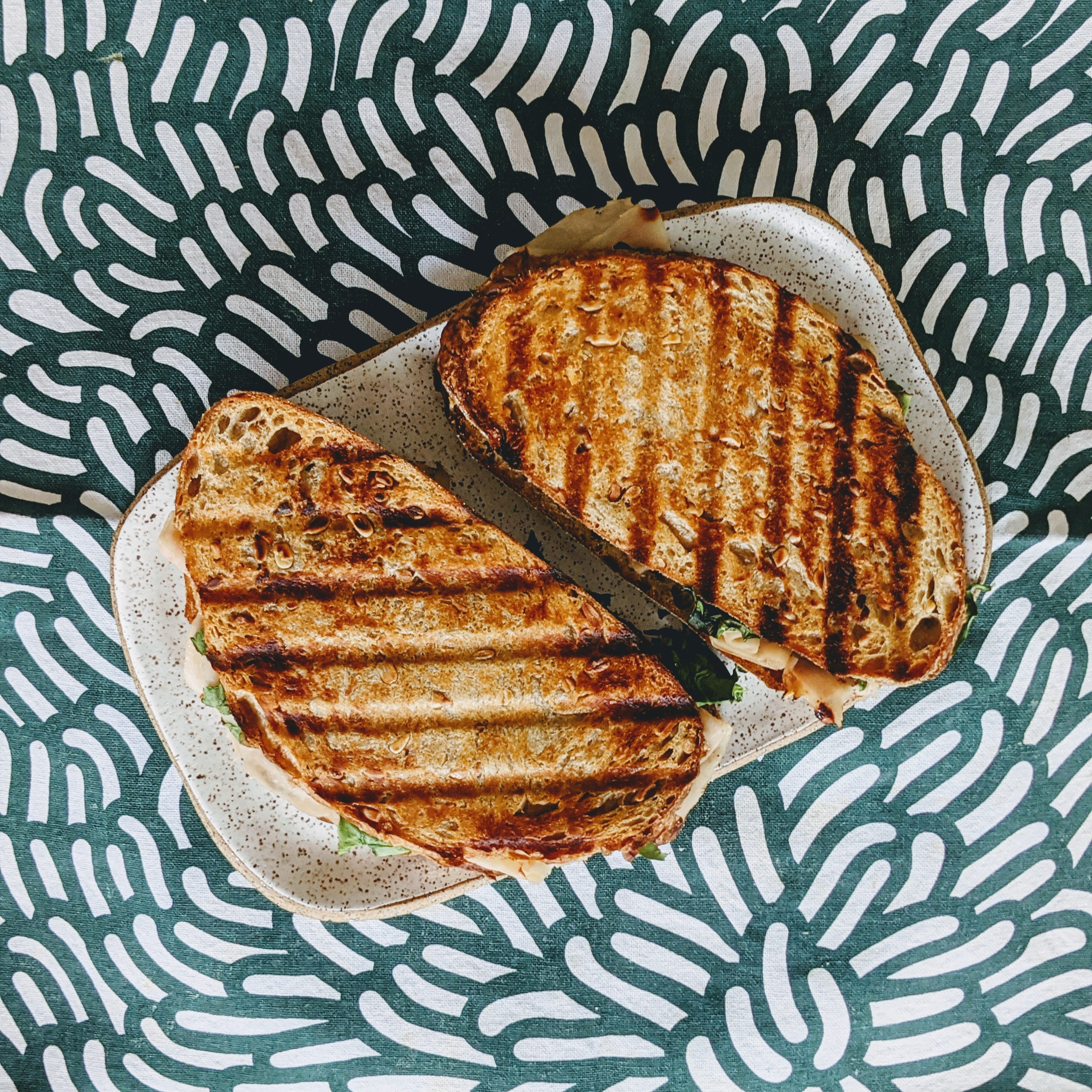 Two grilled panini with diagonal char marks, melted cheese and greens on a speckled plate over teal patterned fabric