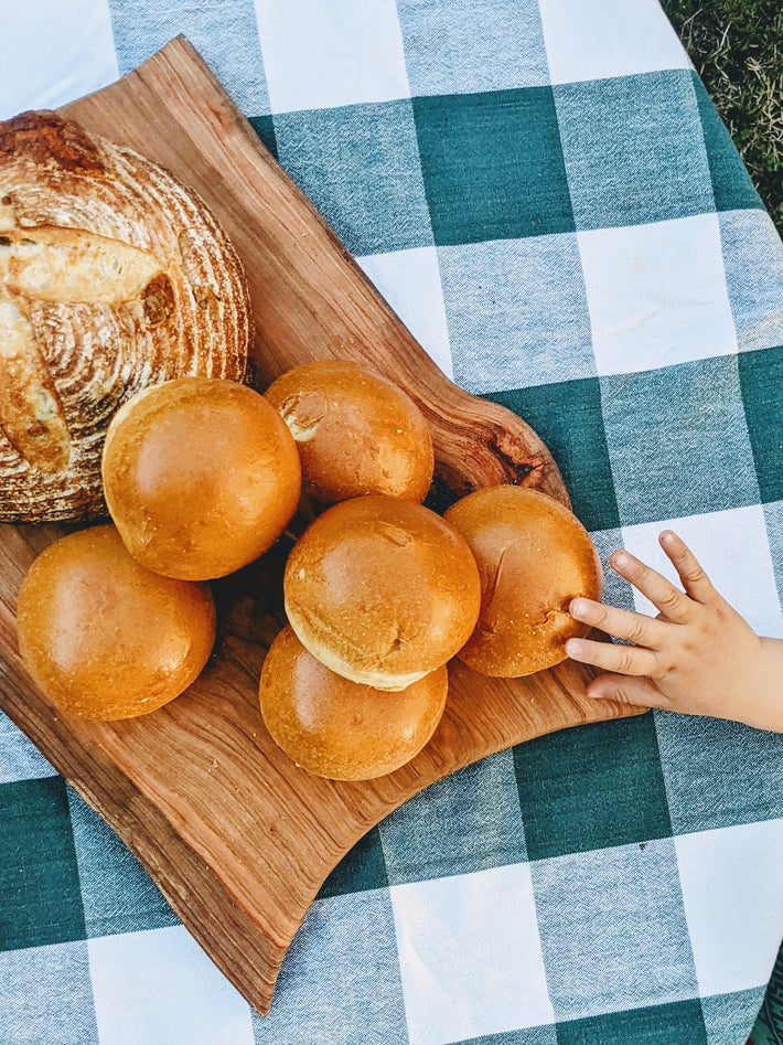 A wooden board with six golden brioche rolls and a round loaf of bread on a checkered tablecloth, with a child's hand reaching for the rolls.