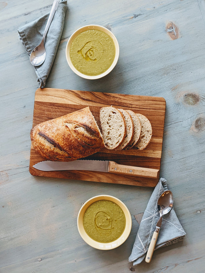 Sliced sourdough bread on a wooden cutting board with two bowls of green soup and utensils