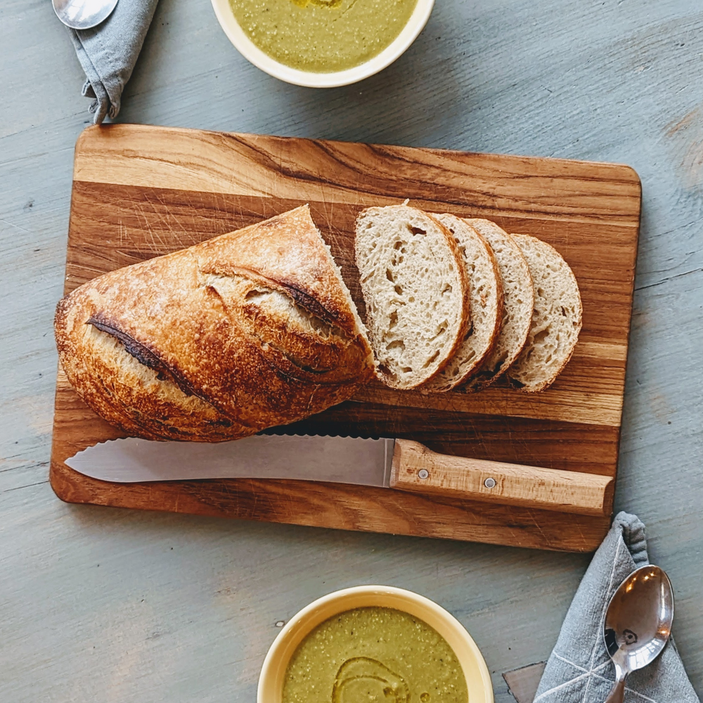 Sliced rustic sourdough loaf on a wooden cutting board with serrated knife; bowls of green soup and a spoon.