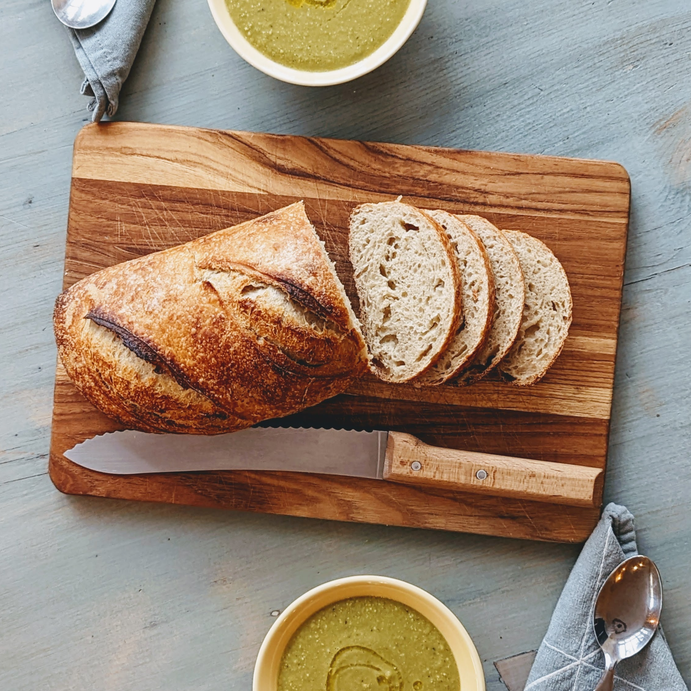 Sliced rustic sourdough loaf on a wooden cutting board with serrated knife; bowls of green soup and a spoon.