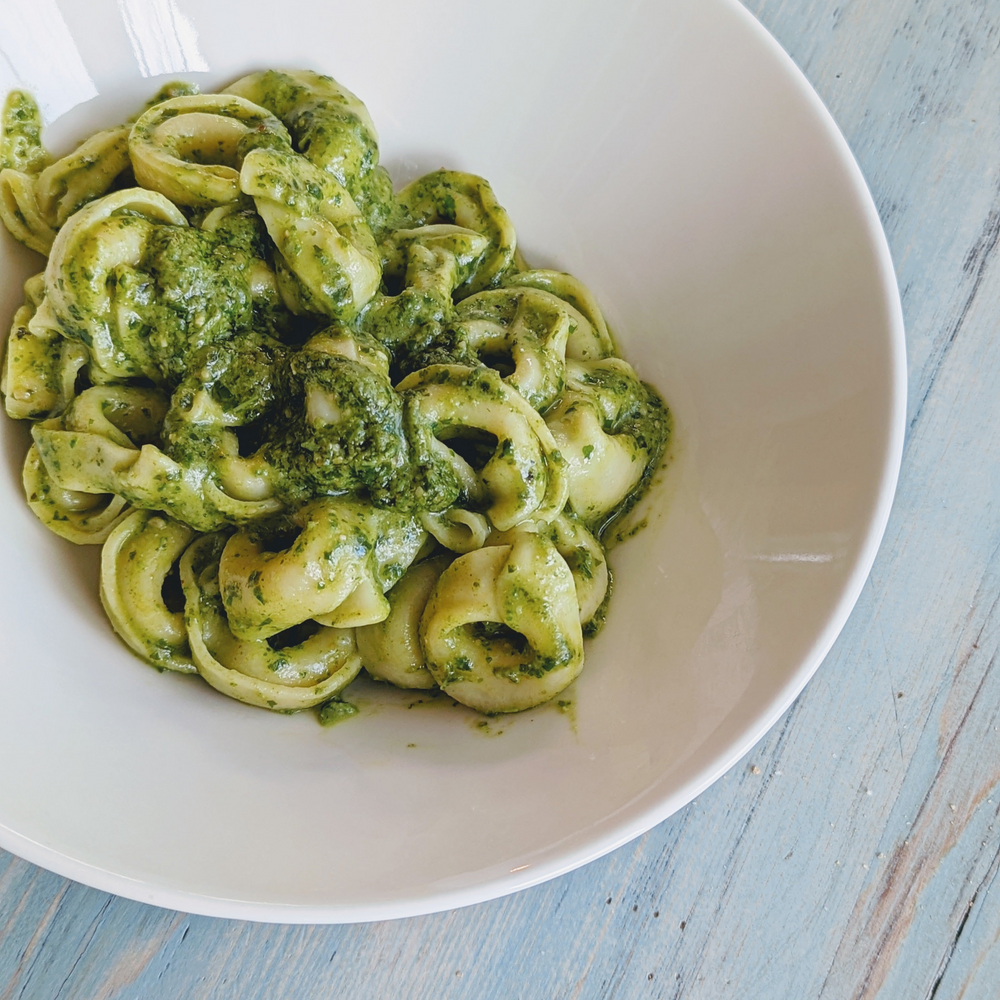 Tortellini pasta coated in green pesto served in a white bowl on a pale blue wooden table