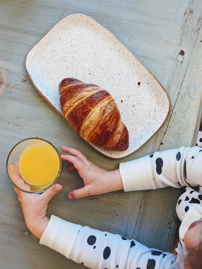 A child's hand holding a glass of orange juice next to a croissant on a plate