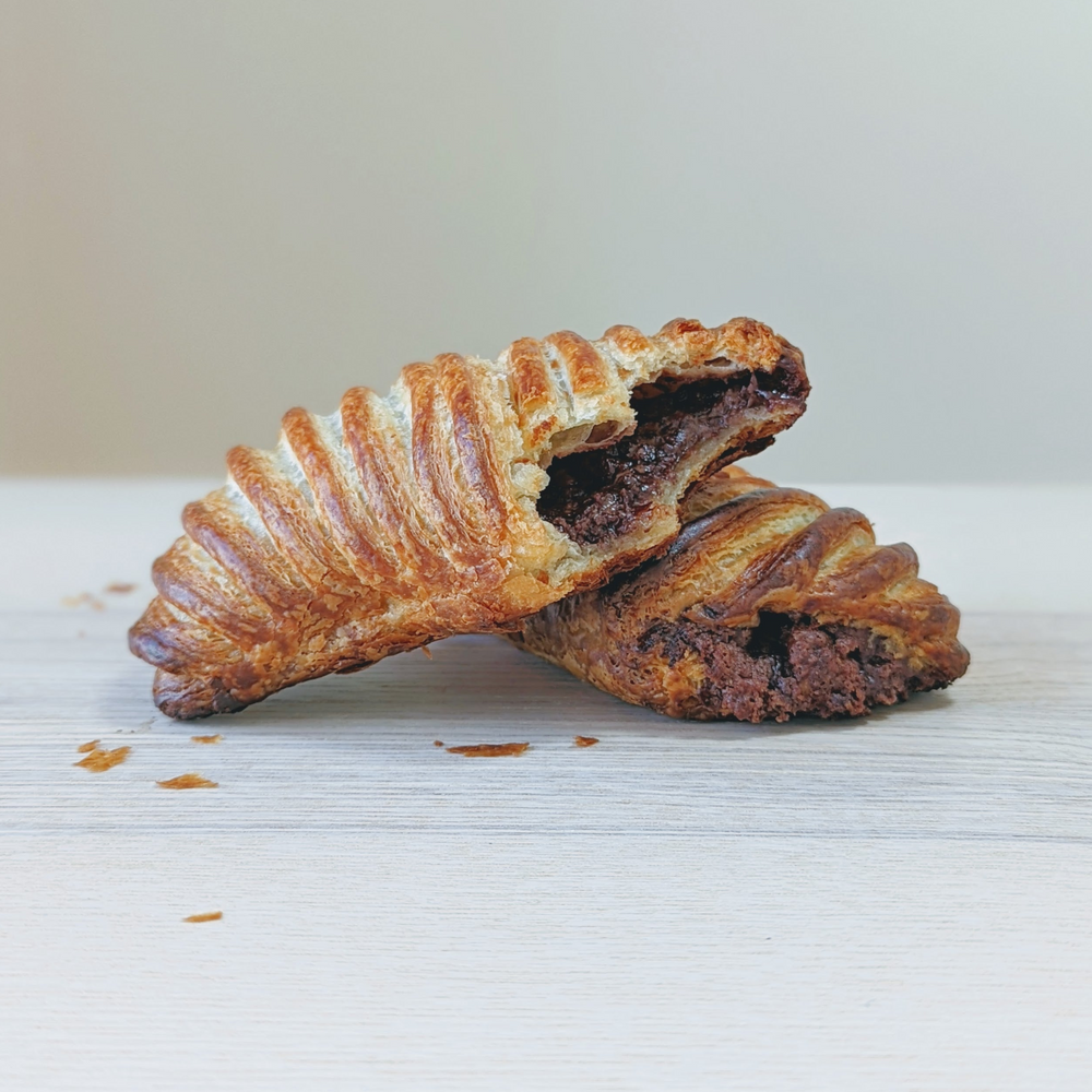 Two chocolate-filled flaky croissants on a light wood surface, one split to show melted chocolate inside.