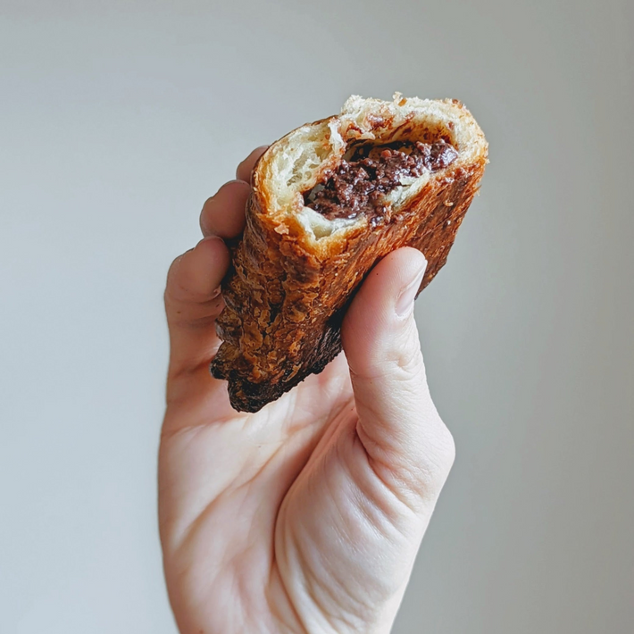 Close-up of Chocolate Avalanche Croissant held in hand showcasing flaky browned crust and melted chocolate filling against a plain gray background