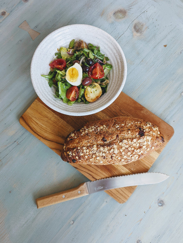 A loaf of bread with oats, a salad with cherry tomatoes and a boiled egg on a wooden cutting board with a knife.