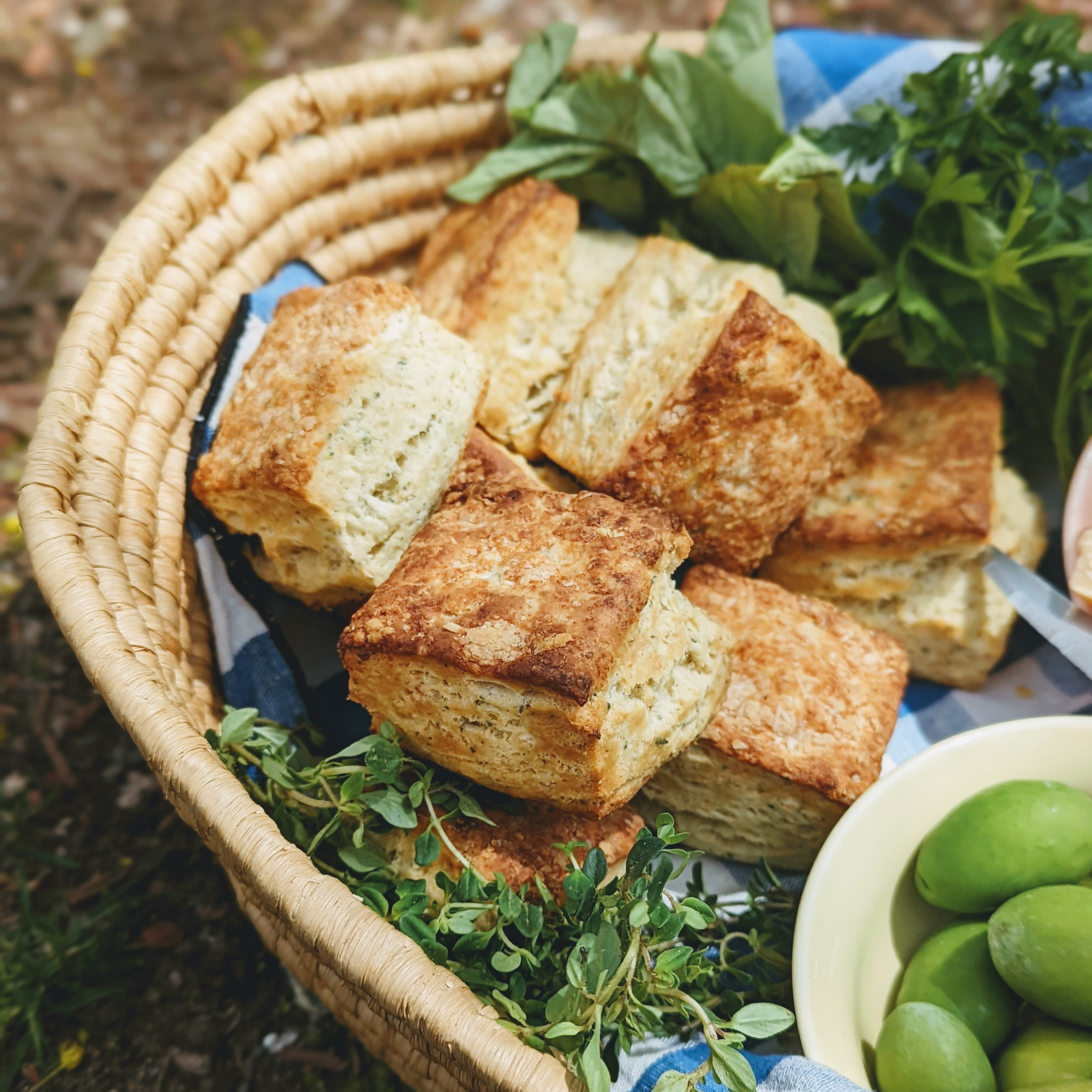 Basket of square herb scones on a cloth with fresh herbs and a small bowl of green olives
