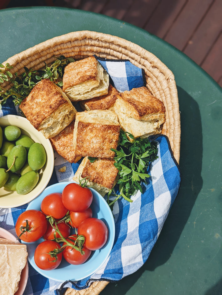 A basket of Parmesan Herb Biscuits, green olives, and tomatoes on a blue checkered cloth.