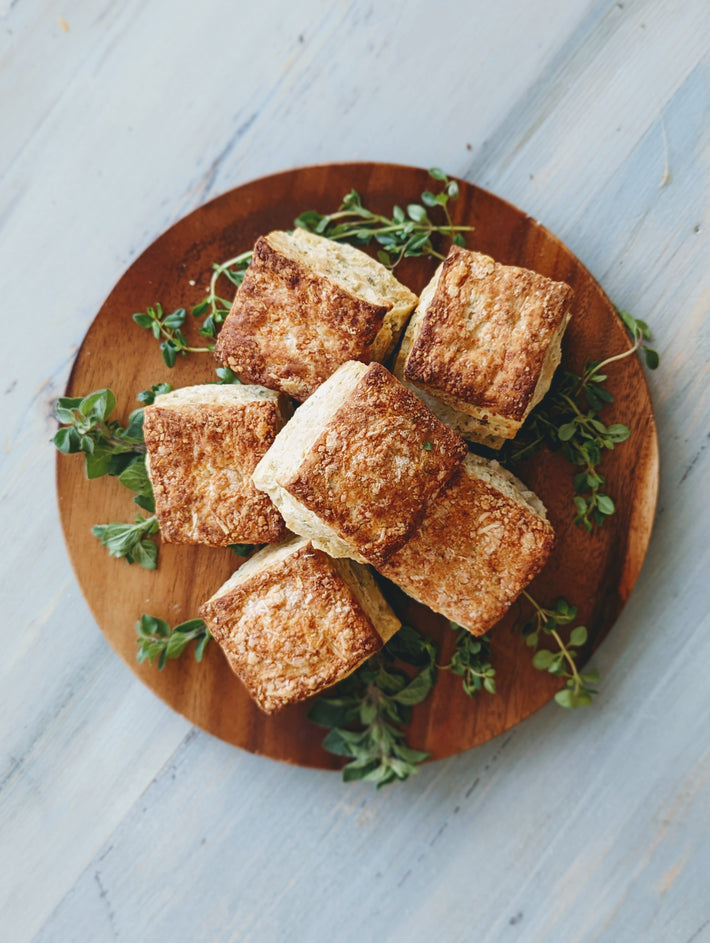 A wooden plate with six golden-brown baked goods surrounded by fresh herbs.