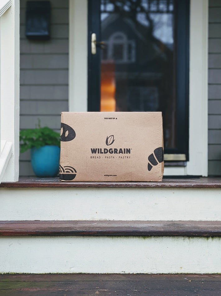 Wildgrain box on front porch step, labeled "THIS WAY UP" with WILDGRAIN, BREAD · PASTA · PASTRY and wildgrain.com