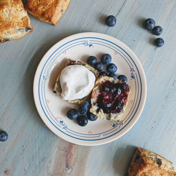 Blueberry scone halves on plate with whipped cream, blueberry jam and scattered blueberries on a light wood table