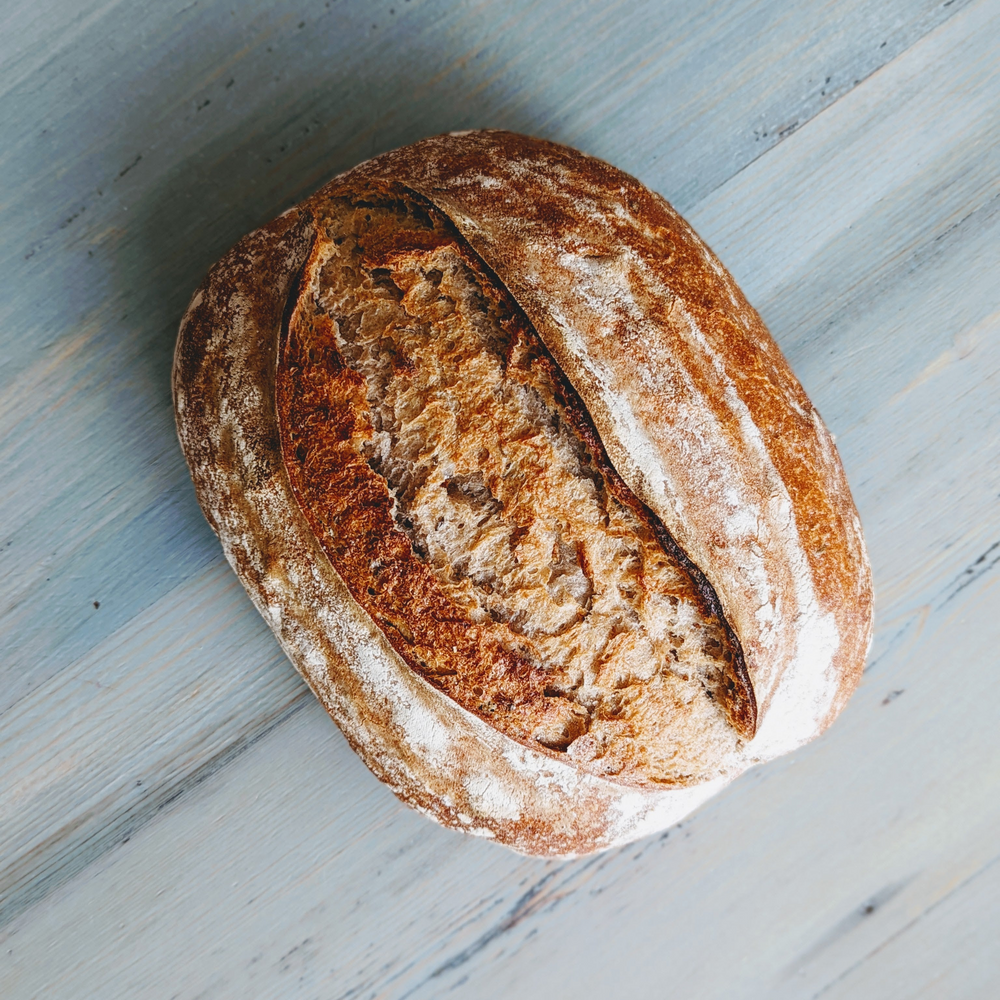 Round crusty sourdough loaf with a central score, dusted with flour on a pale wooden board.