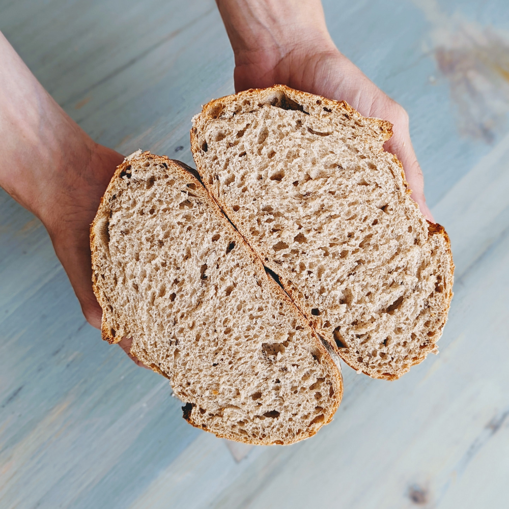 Hands holding a halved rustic whole-grain loaf, showing airy crumb and seeds.