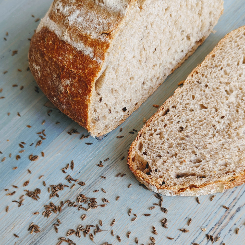 Three-quarter view of sourdough rye loaf with slice showing open crumb and crust, caraway seeds on light blue wood