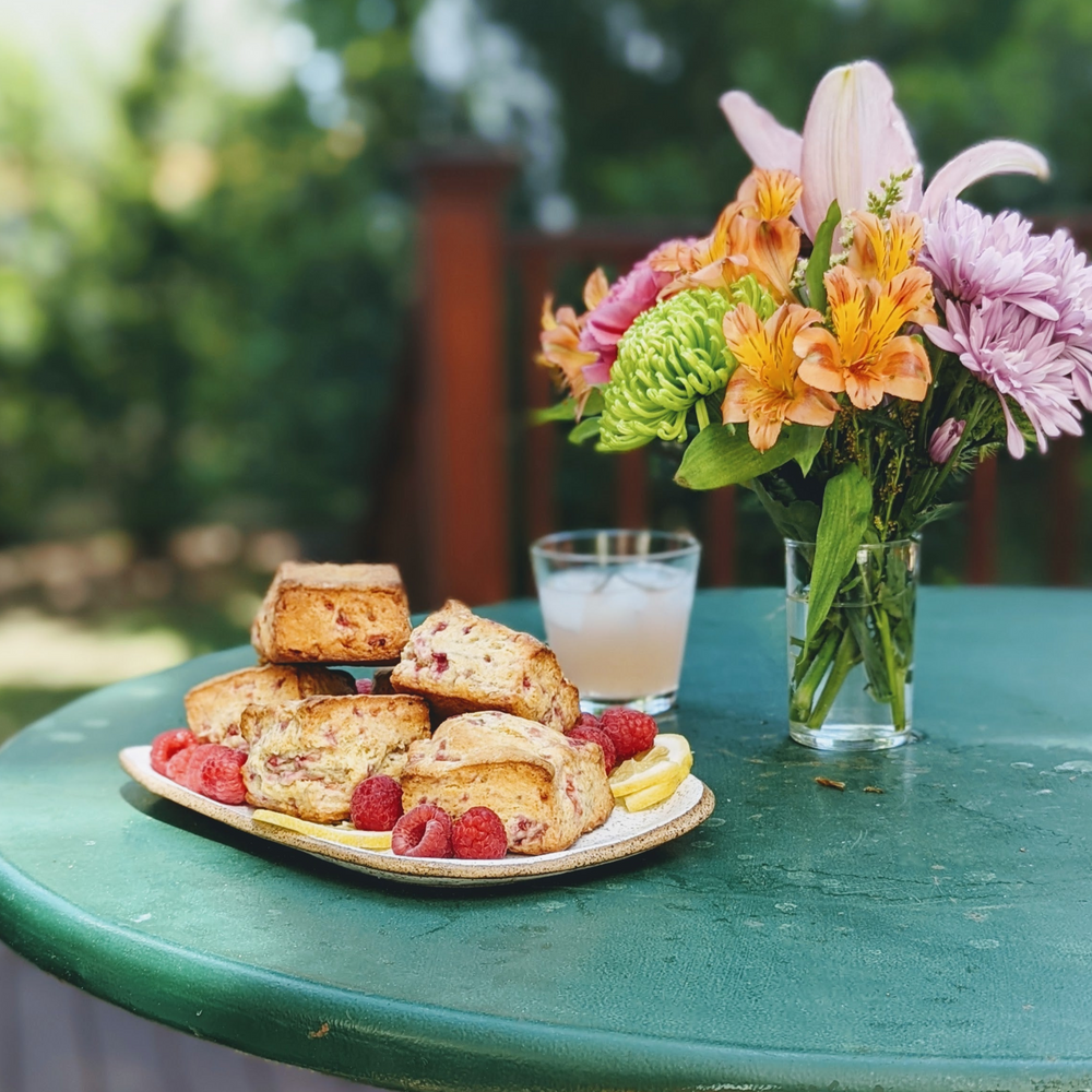 Plate of raspberry scones and lemon slices on a green patio table, with an iced drink and vase of mixed flowers