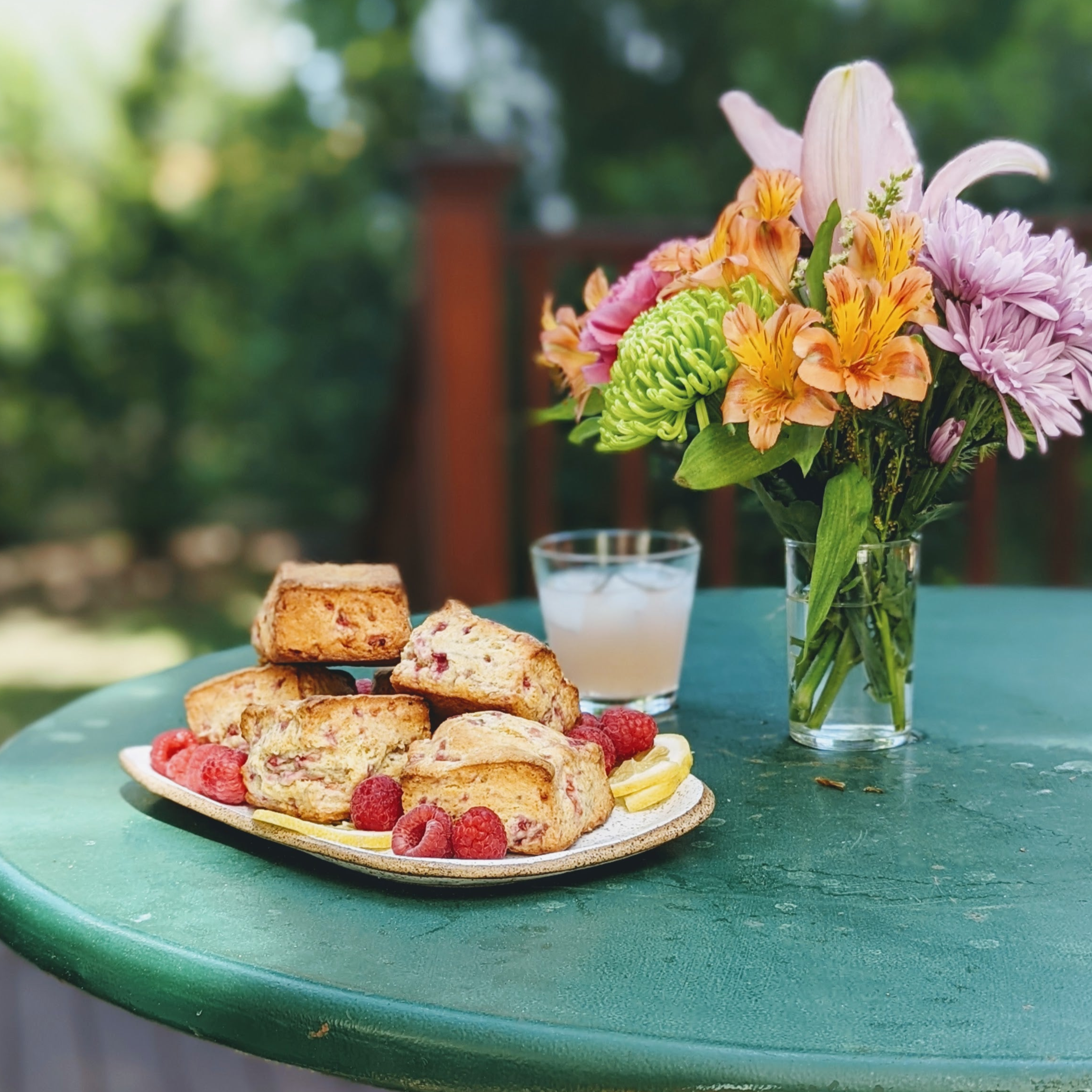 Plate of raspberry scones and lemon slices on a green patio table, with an iced drink and vase of mixed flowers