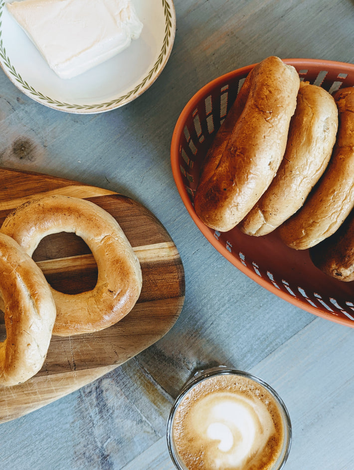 Bagels on a wooden cutting board and in a basket with a plate of cream cheese and a latte