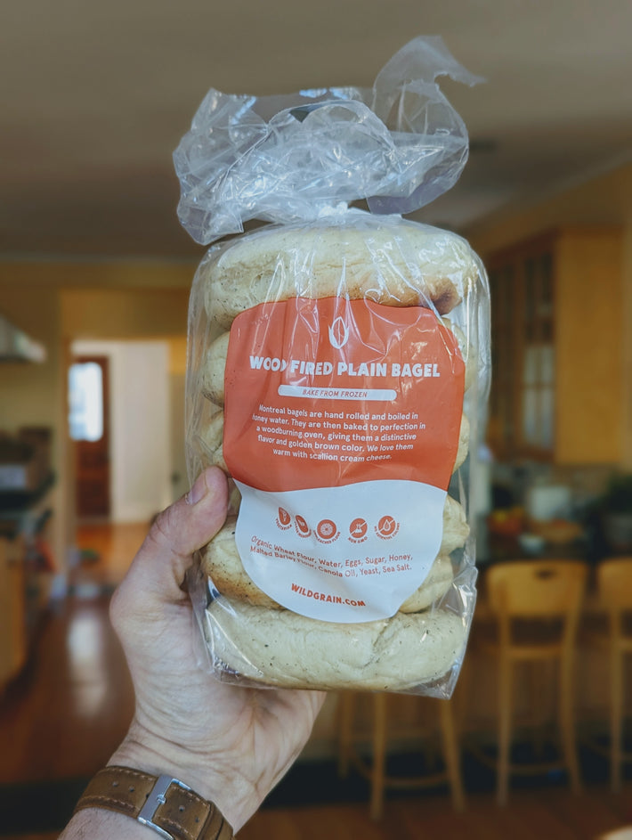 Hand holding Wildgrain bag labeled "WOOD FIRED PLAIN BAGEL" with visible "BAKE FROM FROZEN" and WILDGRAIN.COM