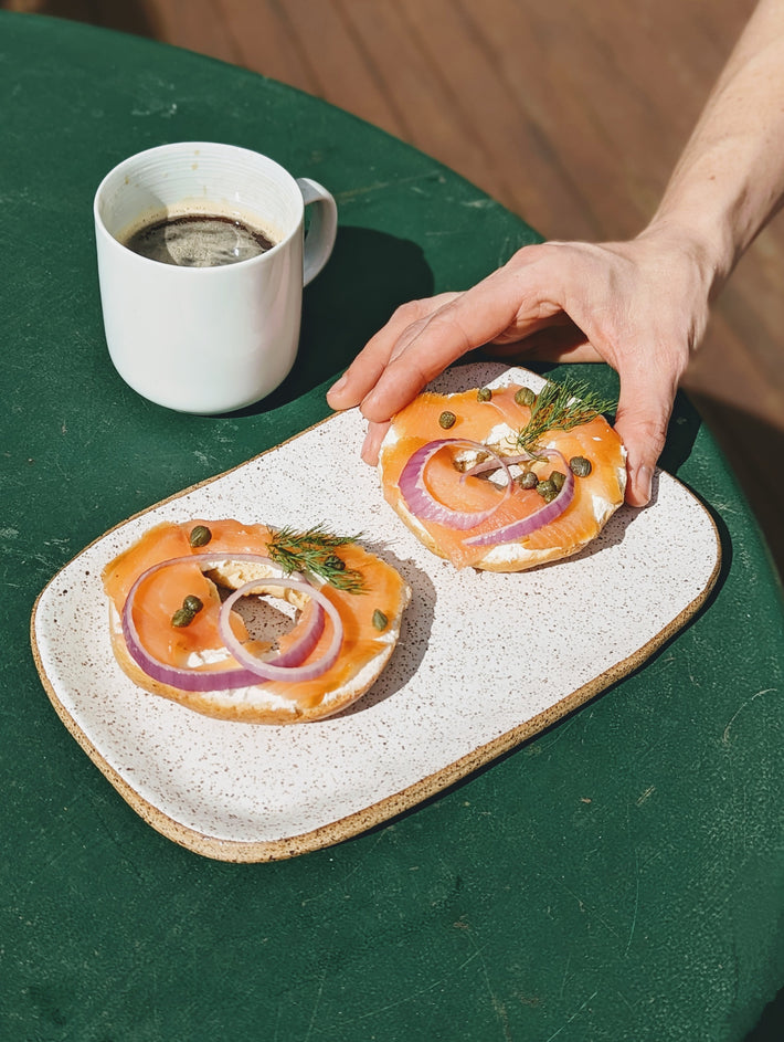 A hand holding a plate with two bagels topped with smoked salmon, red onion, and capers, next to a cup of coffee.