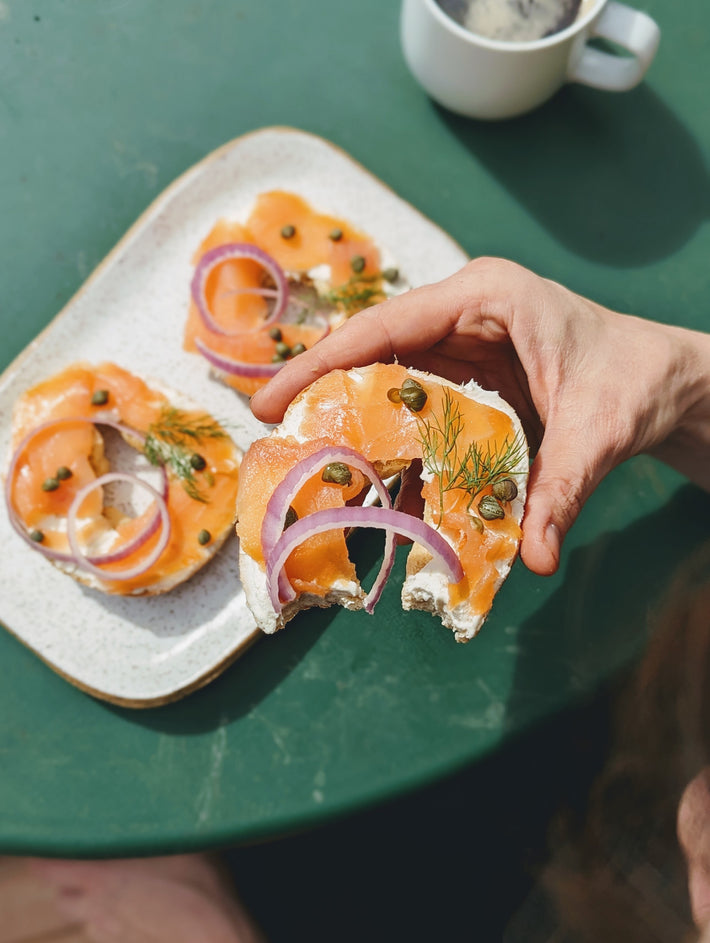 Hand holding smoked-salmon bagel half with cream cheese, red onion, capers and dill; plate and coffee in background