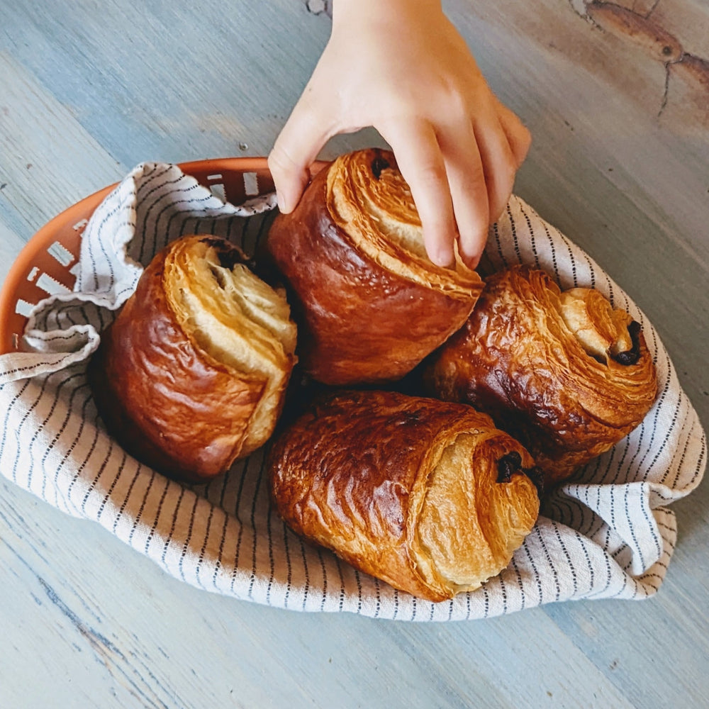 Four pain au chocolat in a striped cloth-lined basket, one pastry being reached for by a child's hand.