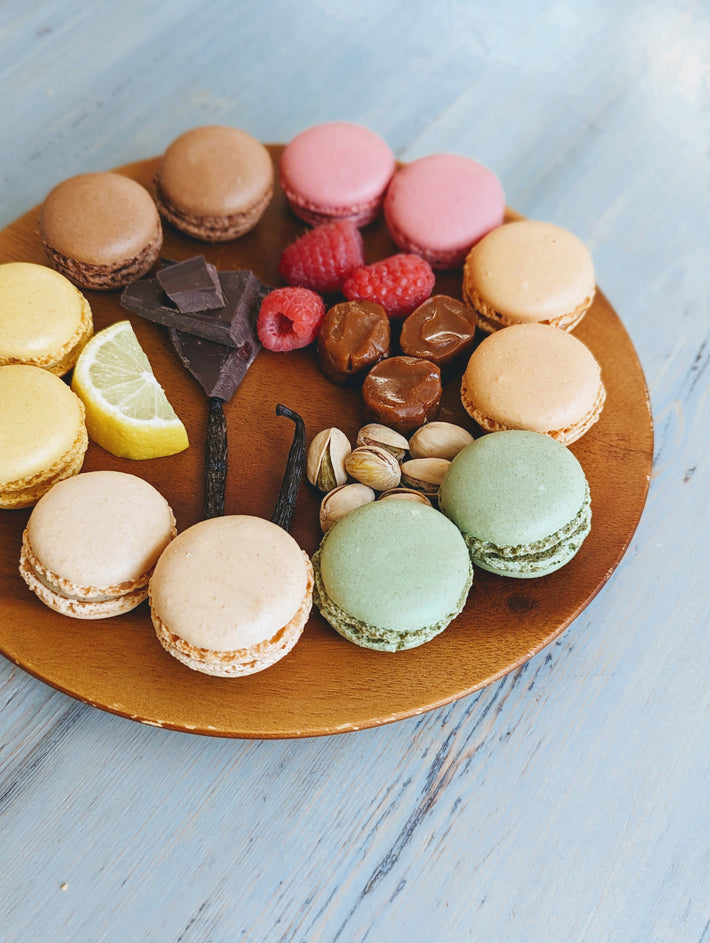 A colorful assortment of macarons on a wooden platter, including flavors like lemon, pistachio, and raspberry, alongside chocolate and nuts.