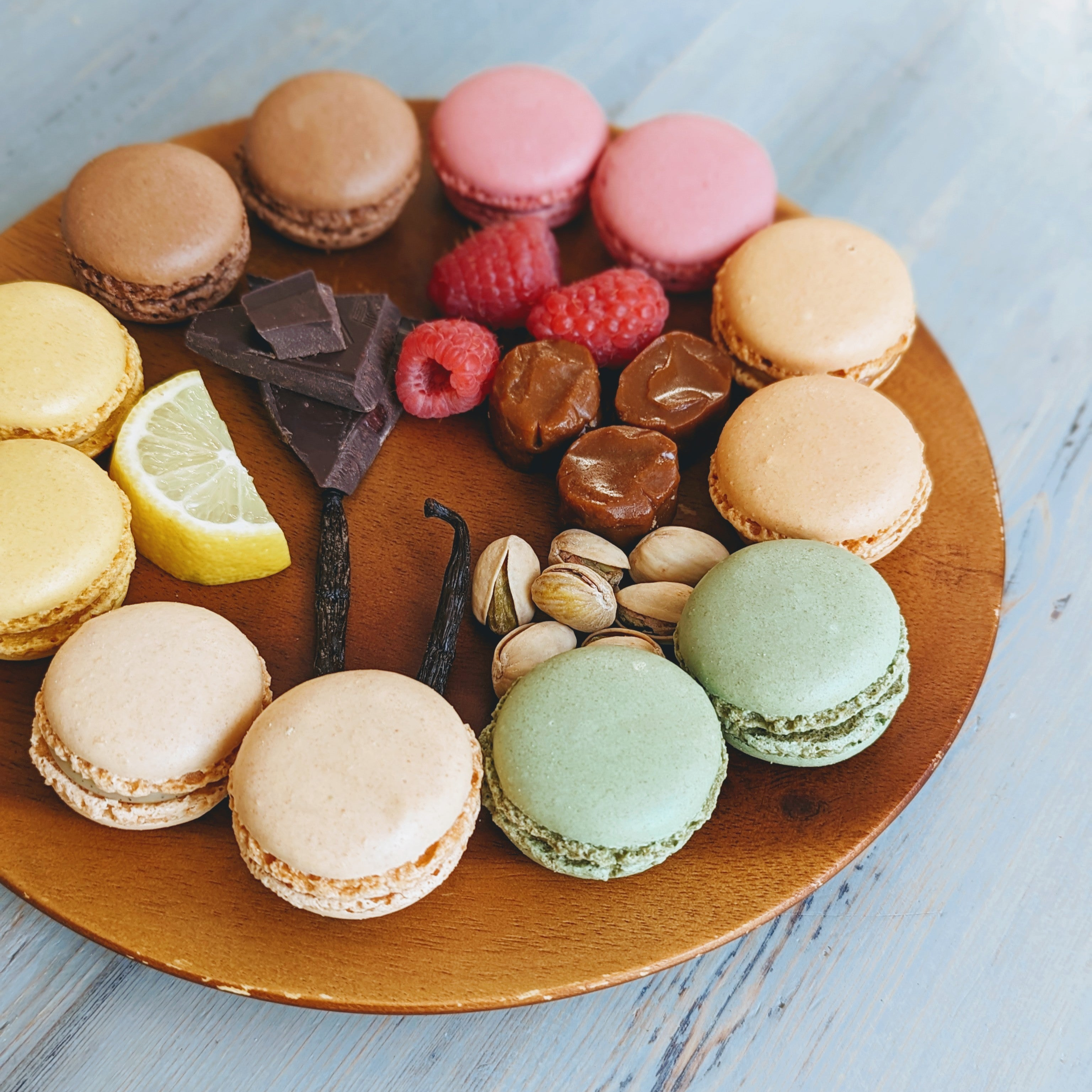 Assorted pastel macarons arranged on a wooden board with chocolate pieces, raspberries, pistachios and a lemon wedge.
