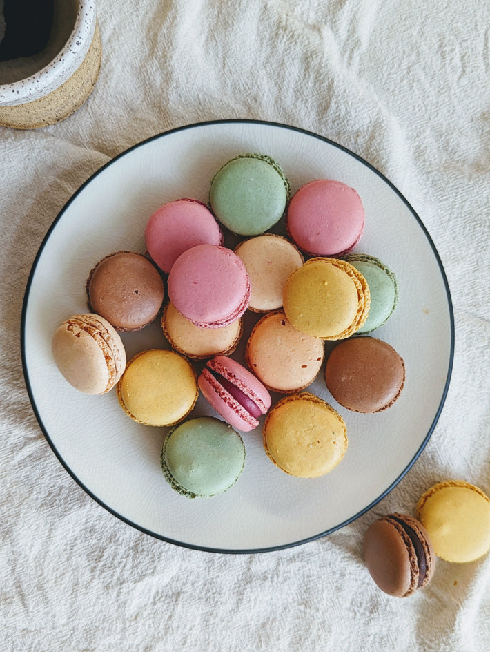 Assorted colorful macarons on a white plate with a dark rim, placed on a light cloth surface next to a cup.
