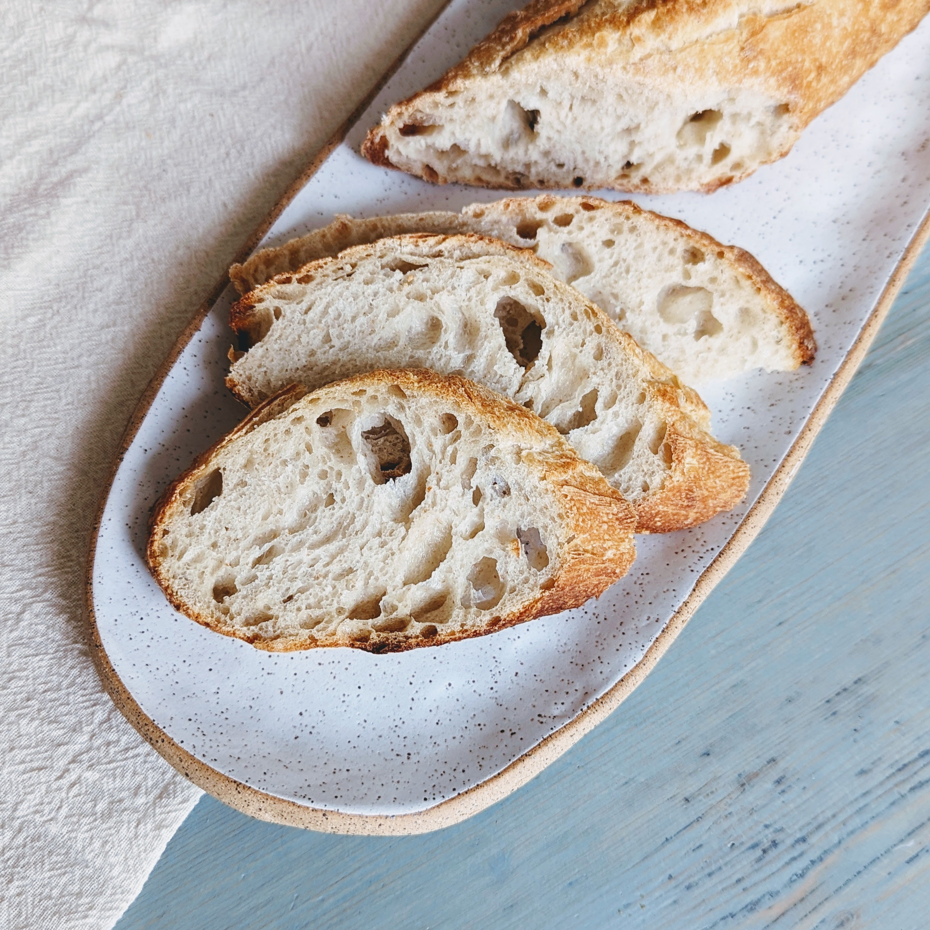 Sliced crusty baguette on a speckled oval plate beside a light cloth on a blue wooden table.