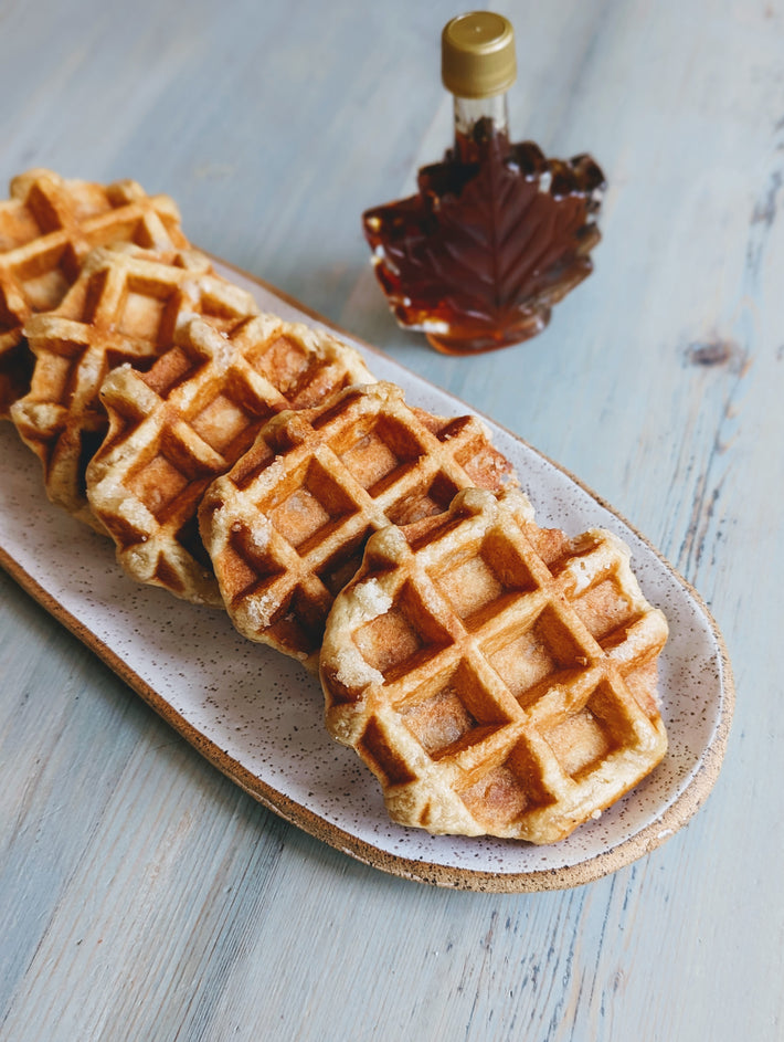 Five artisanal waffles on a plate with a bottle of maple syrup in the background.