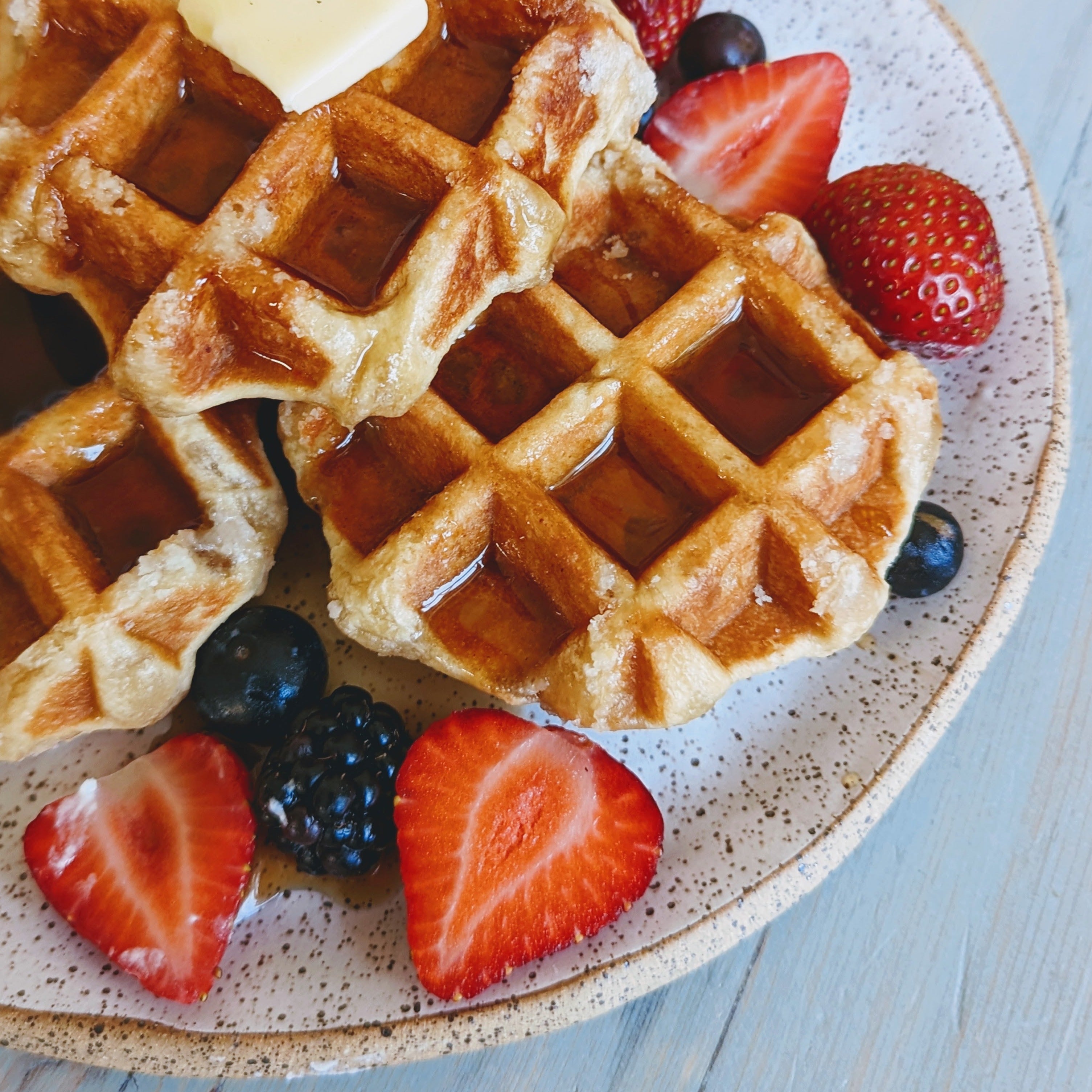 Belgian-style waffles topped with butter and syrup on a speckled plate with strawberries, blueberries and blackberries.