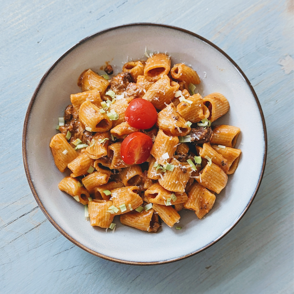 Bowl of rigatoni in creamy tomato sauce topped with cherry tomatoes, Parmesan and chives on light blue wood.
