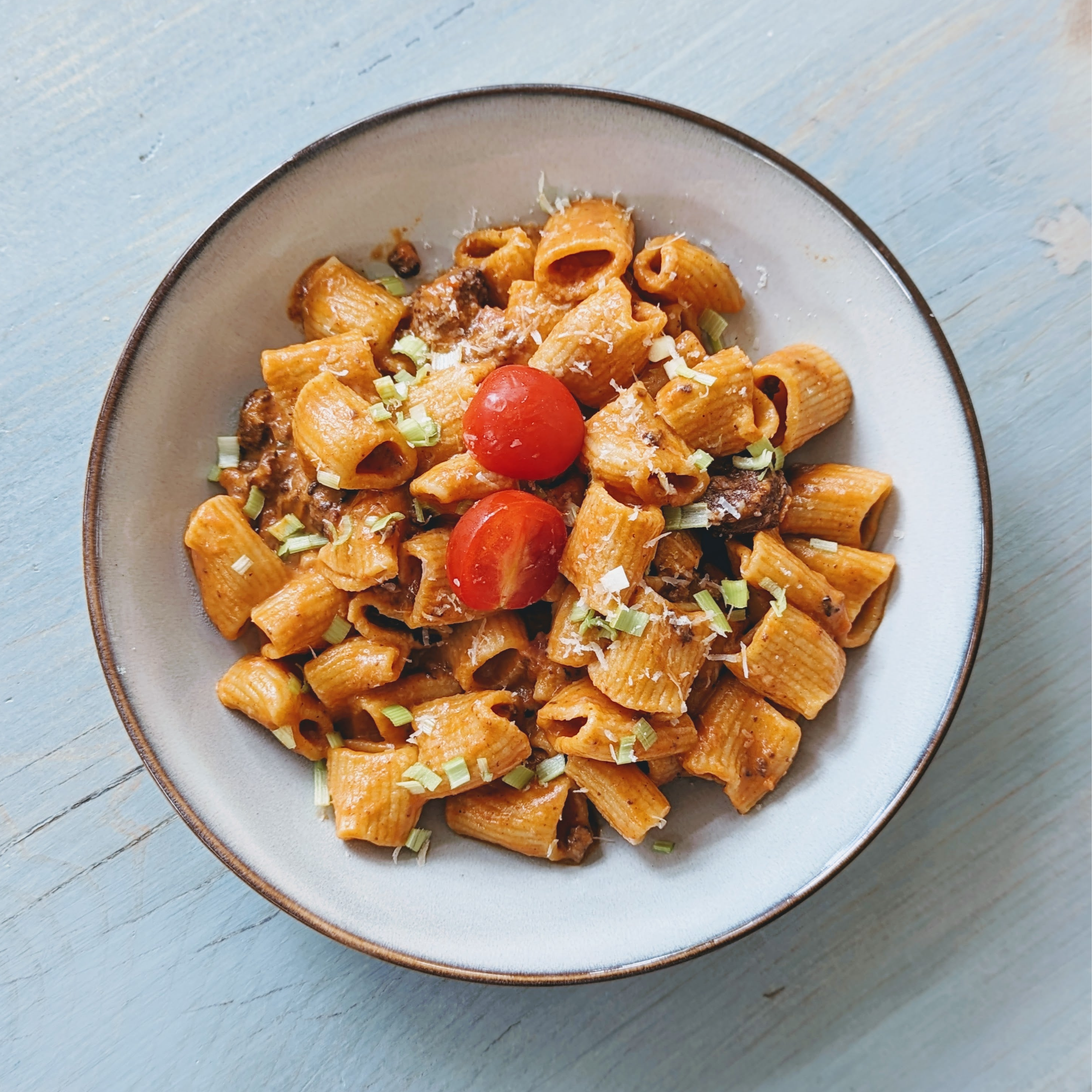Bowl of rigatoni in creamy tomato sauce topped with cherry tomatoes, Parmesan and chives on light blue wood.
