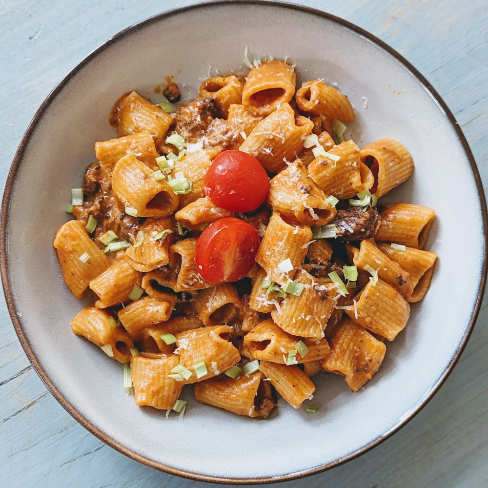 Short ridged pasta in creamy tomato sauce with halved cherry tomatoes and chopped chives in white bowl on a light blue table