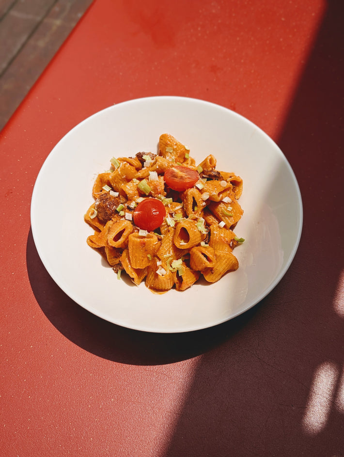 White bowl of tomato pasta with cherry tomatoes and chopped scallions on a red table lit by sunlight and shadow.