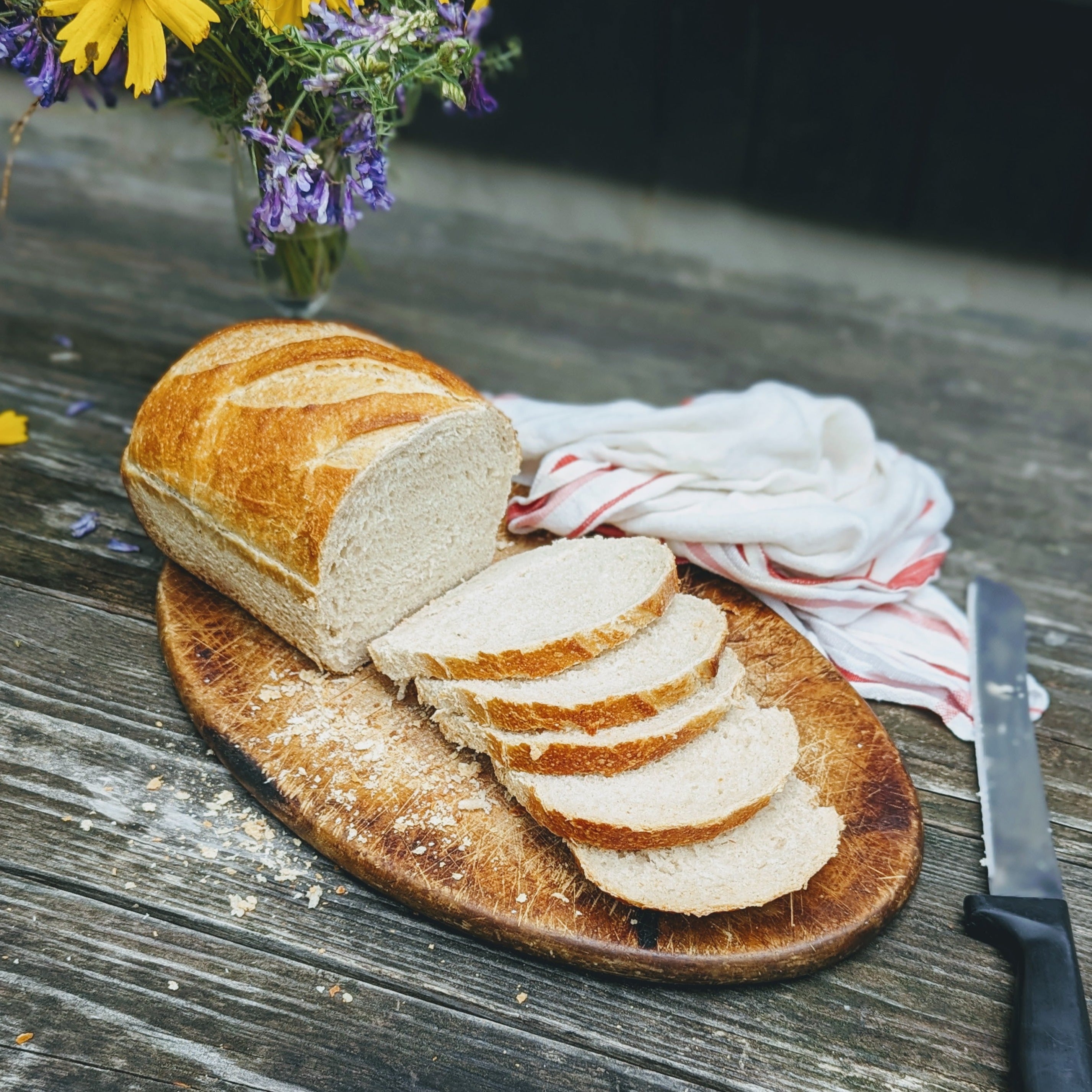 Sliced sourdough sandwich loaf on a wooden board with a knife and flowers in the background