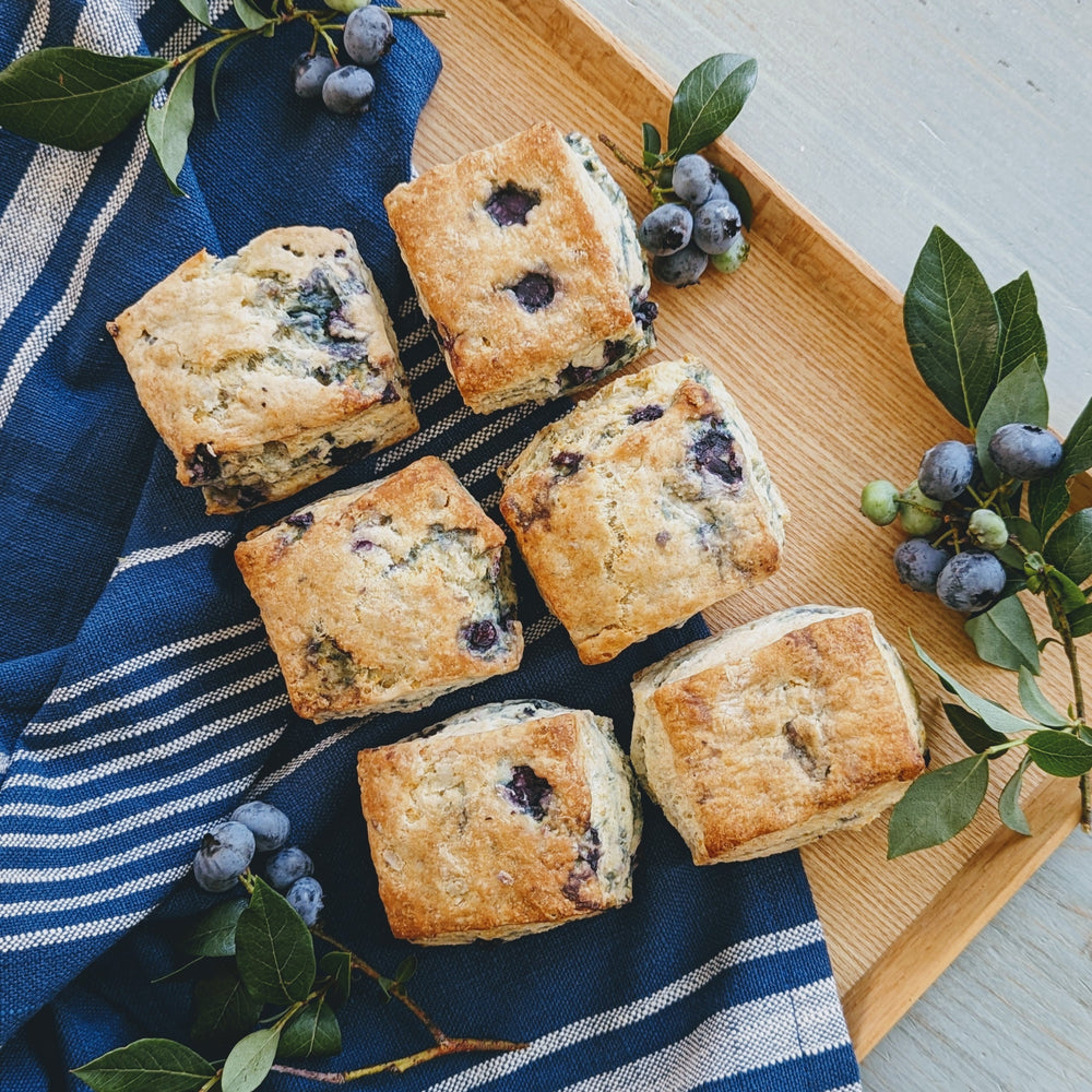 Six plant-based blueberry biscuits arranged on a wooden tray with blueberries and leaves.