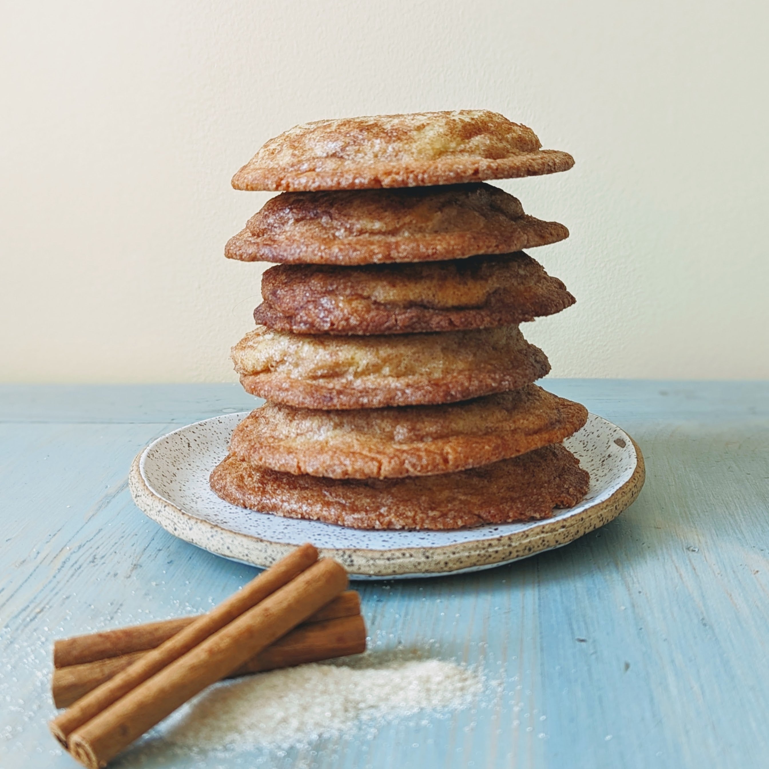Stack of six golden-brown cookies on a ceramic plate with cinnamon sticks and a dusting of sugar on blue wooden table.