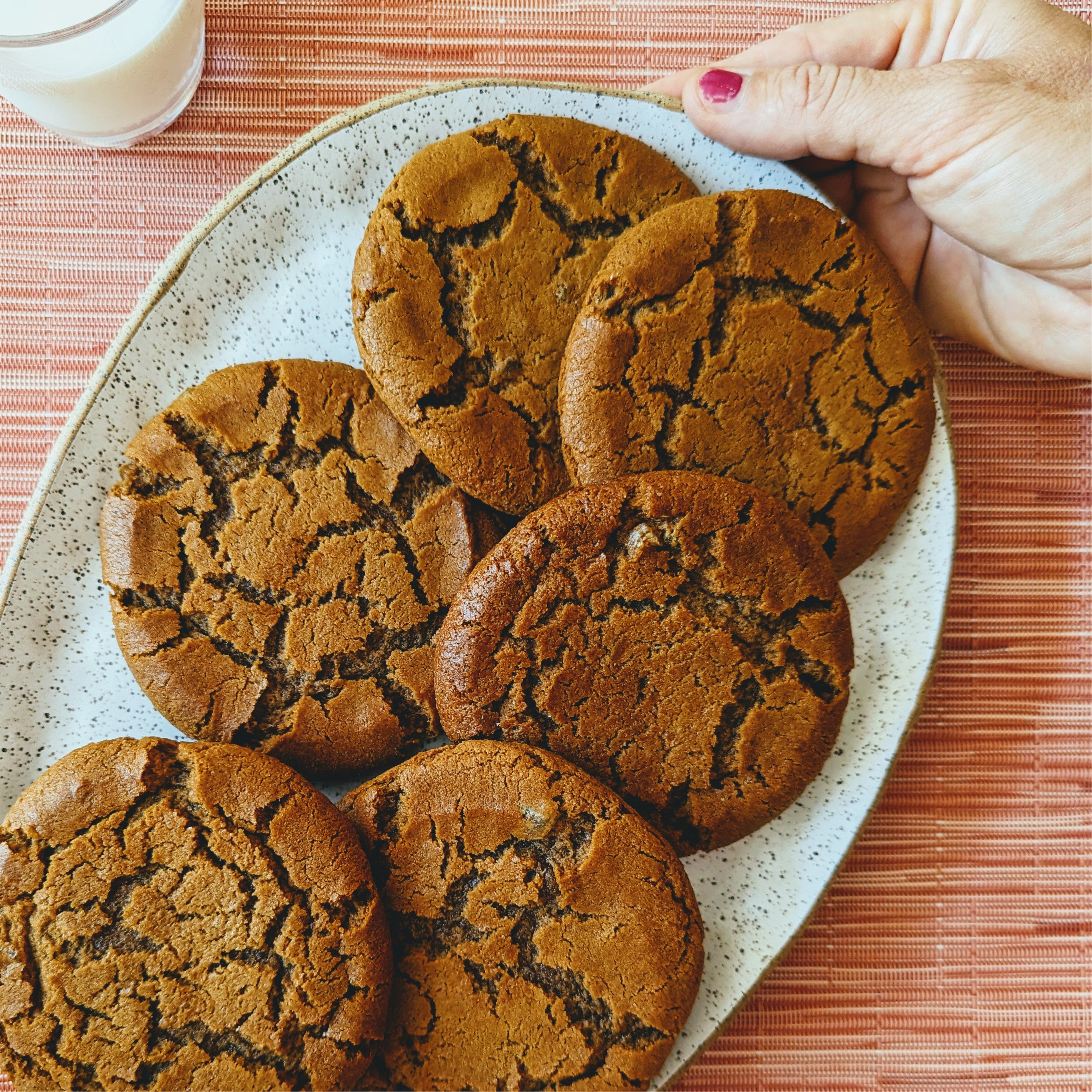 Six cracked-top ginger molasses cookies on a speckled plate, hand reaching from top right; glass of milk at upper left