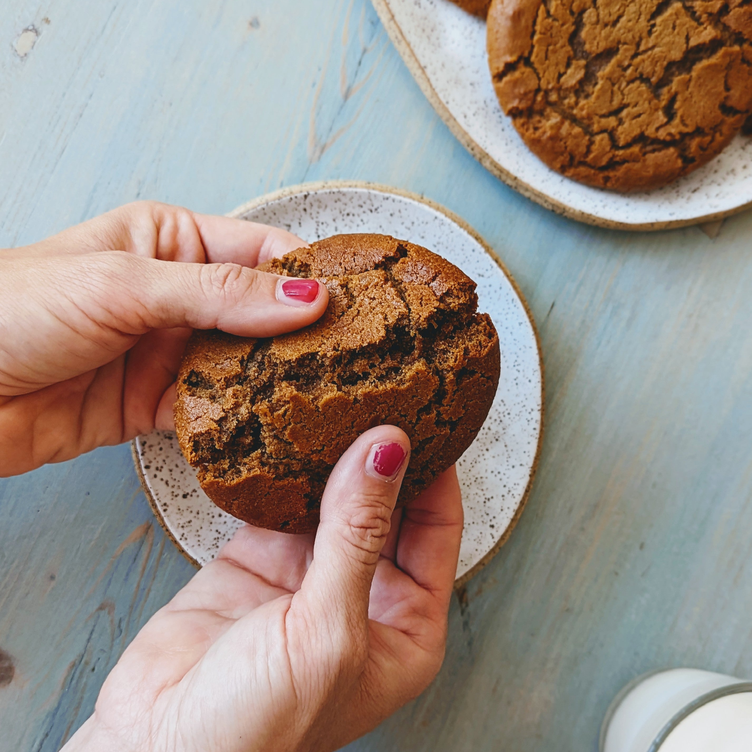 Hands breaking a large cracked chocolate cookie over a speckled plate on a blue wooden table, with extra cookies and a glass of milk nearby.
