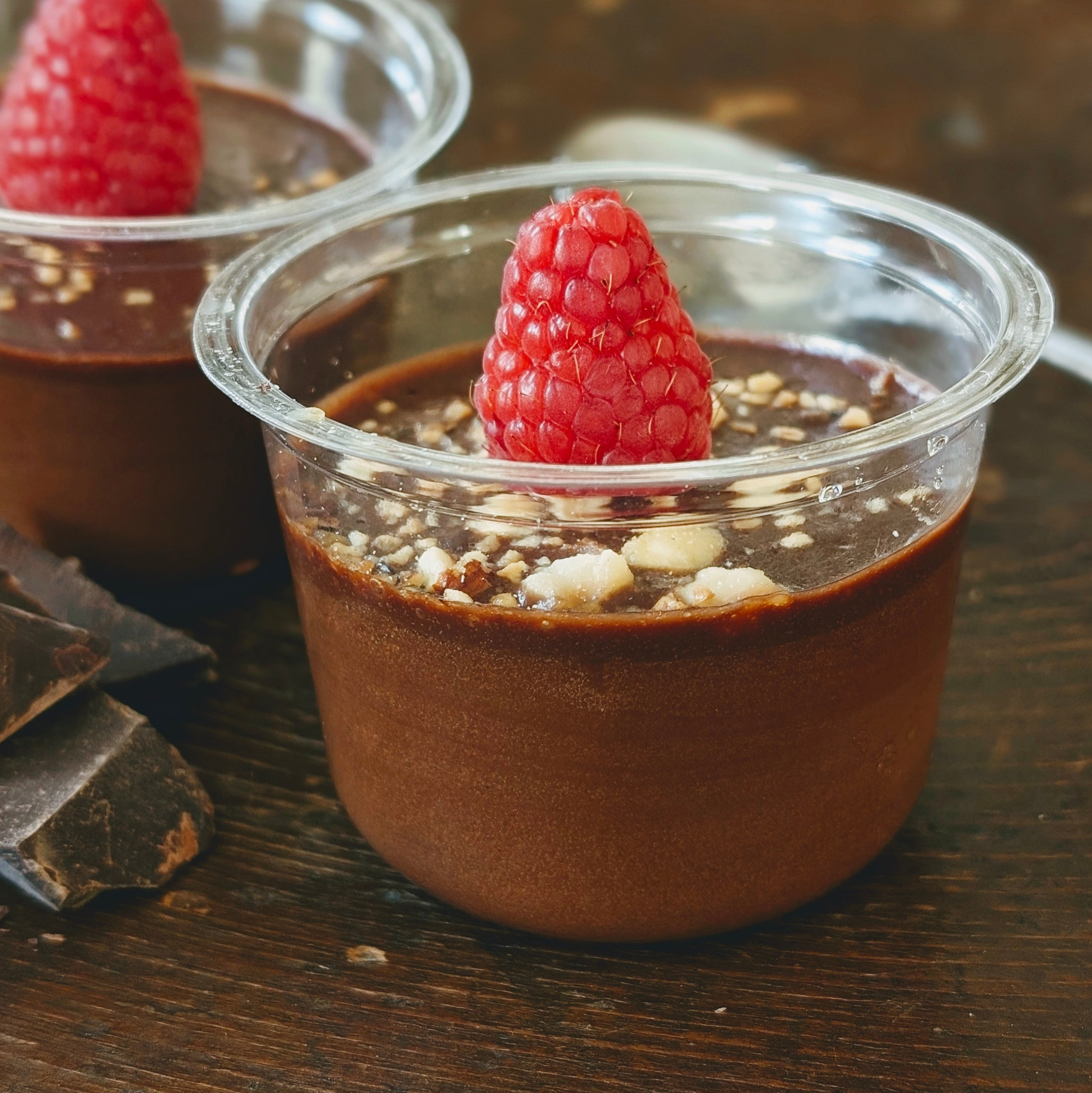 Chocolate mousse in a small clear cup topped with a raspberry and chopped nuts, with chocolate pieces nearby
