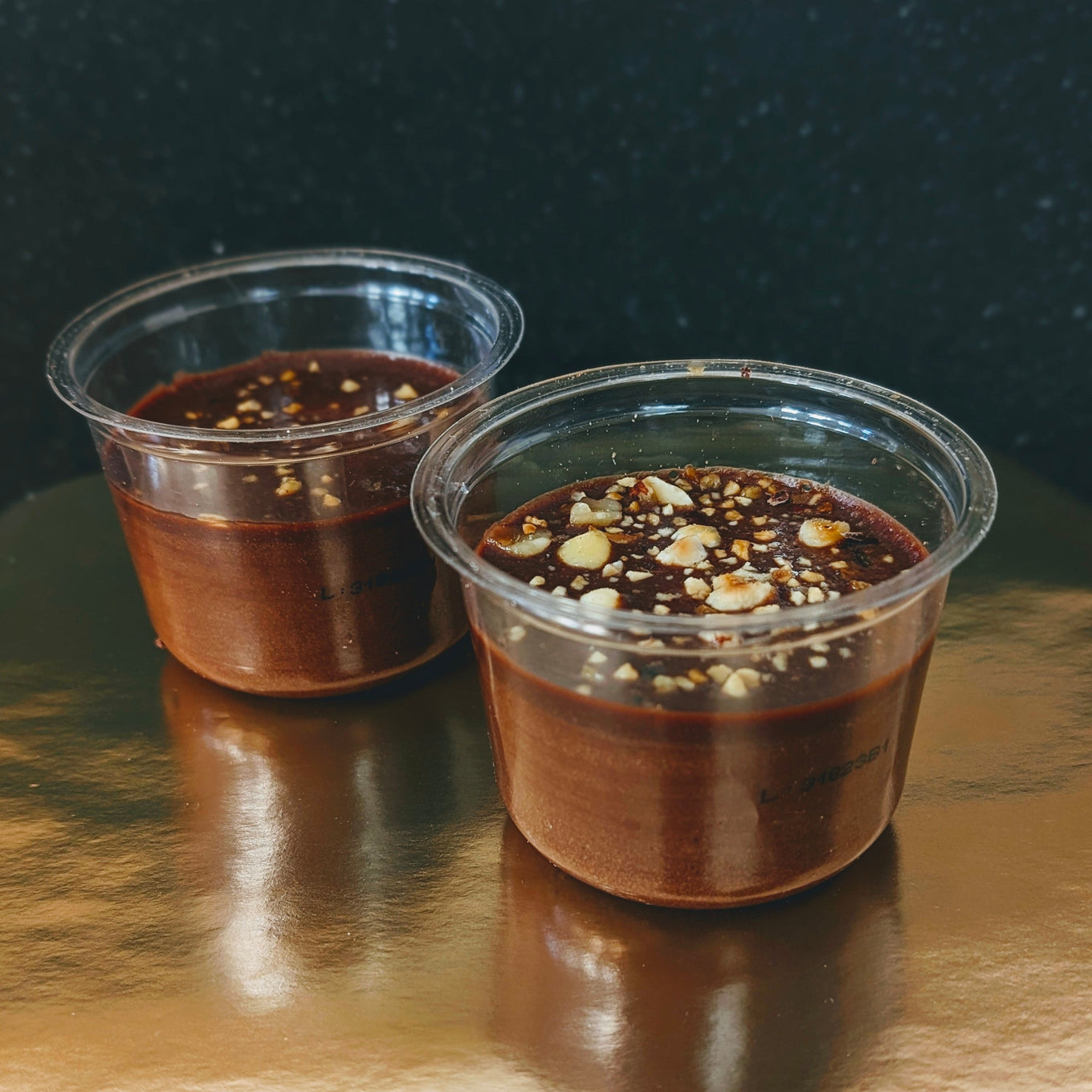 Two clear plastic cups filled with chocolate dessert topped with chopped nuts on a reflective surface.